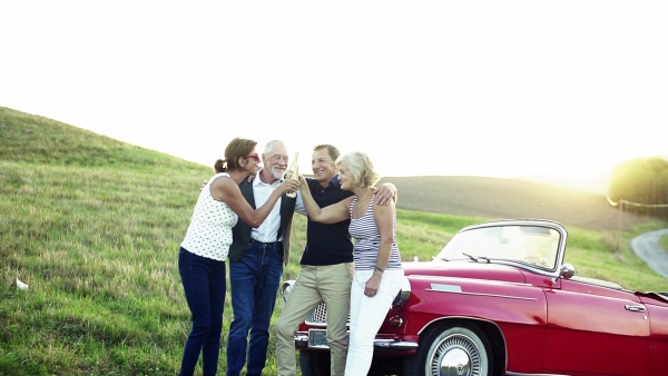 Two senior couples standing by cabriolet on a road trip in summer, clinking bottles. Slow motion.