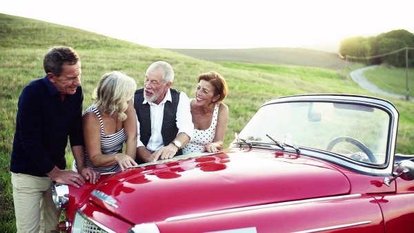 Two senior couples standing by cabriolet on a road trip in summer, looking at the map and planning.