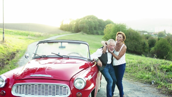 A cheerful senior couple in love standing by cabriolet on a road trip in summer at sunset, hugging.