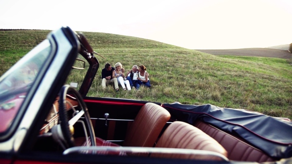 Two senior couples sitting by cabriolet on a road trip in summer, talking. Slow motion.