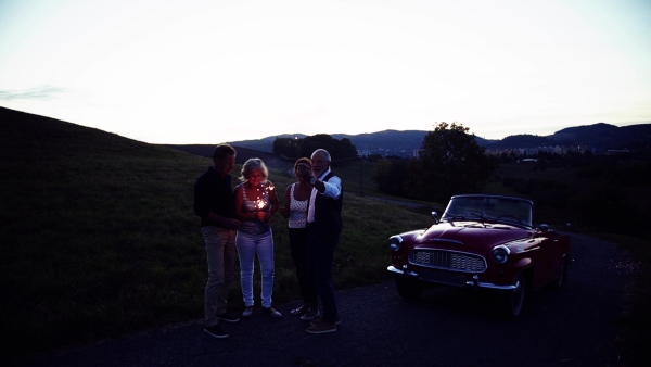 Two senior couples with sparklers standing by cabriolet on a road trip in summer, having fun. Slow motion.