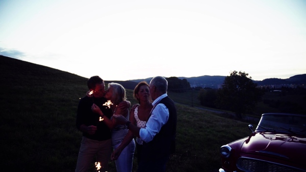 Two senior couples with sparklers standing by cabriolet on a road trip in summer, having fun. Slow motion.