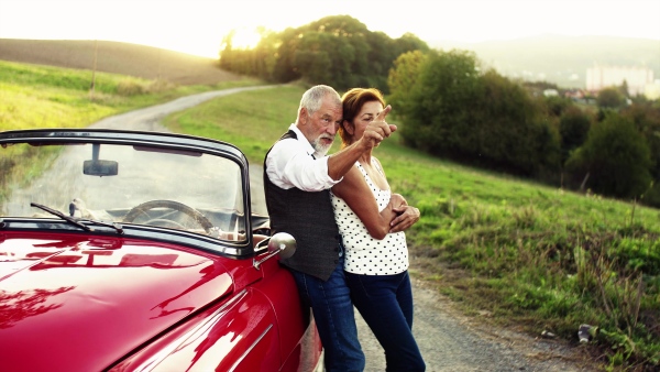 A cheerful senior couple in love standing by cabriolet on a road trip in summer at sunset, talking.