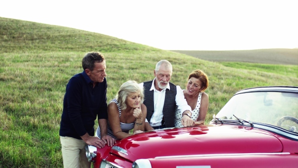 Two senior couples standing by cabriolet on a road trip in summer, looking at the map and planning.