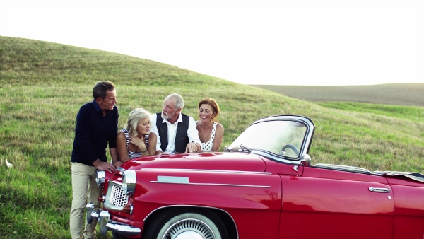 Two senior couples standing by cabriolet on a road trip in summer, looking at the map and planning.