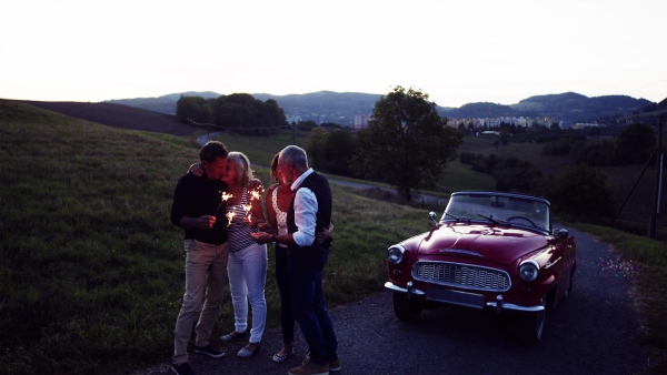 Two senior couples with sparklers standing by cabriolet on a road trip in summer at dusk, having fun.