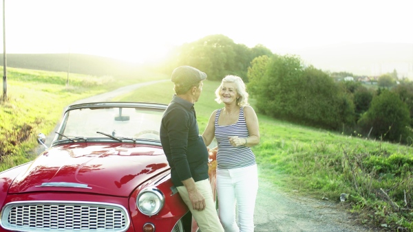 A cheerful senior couple in love standing by cabriolet on a road trip in summer at sunset, hugging.