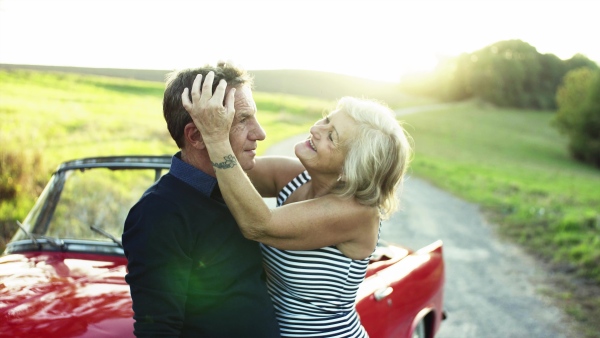 A cheerful senior couple in love standing by cabriolet on a road trip in summer at sunset, hugging.