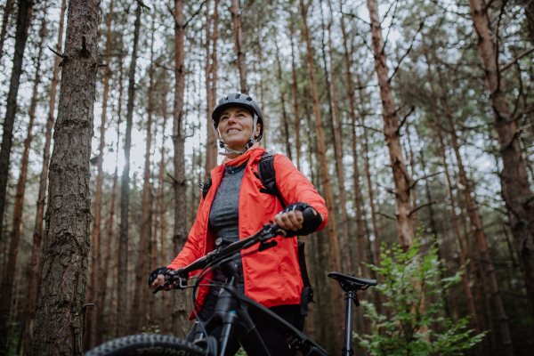 A low angle view of senior woman biker walking and pushing bike outdoors in forest in autumn day.