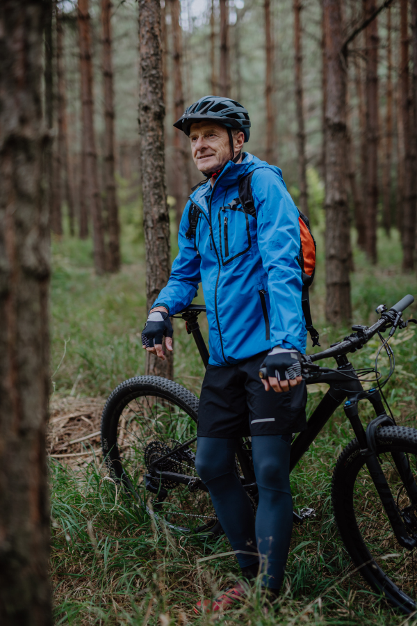 A n active senior man biker standing with bike outdoors in forest in autumn day.