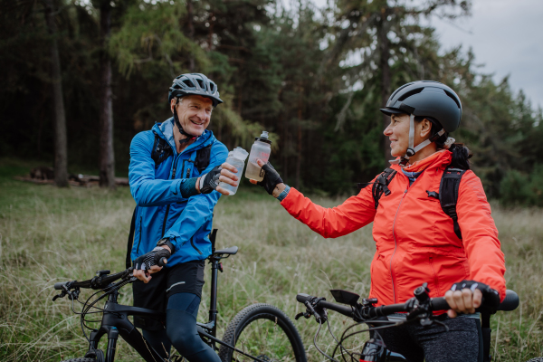 A happy senior couple bikers with water bottles outdoors in forest in autumn day.