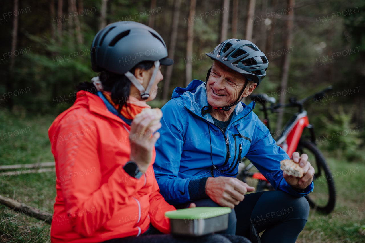 A happy senior couple bikers with eating snack outdoors in forest in autumn day.