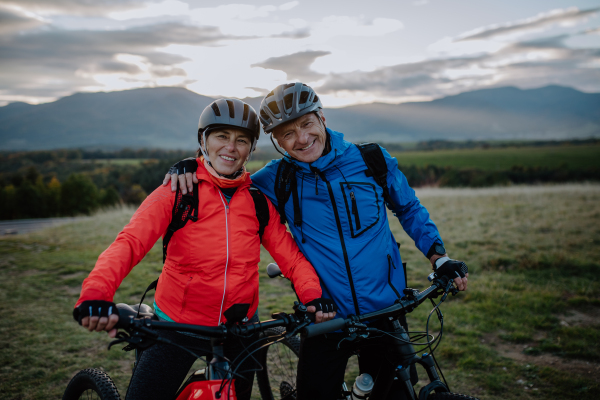 A happy senior couple bikers embracing outdoors in nature in autumn day, looking at camera.