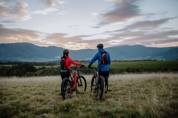 A rear view of senior couple bikers walking and pushing e-bikes outdoors in forest in autumn day.