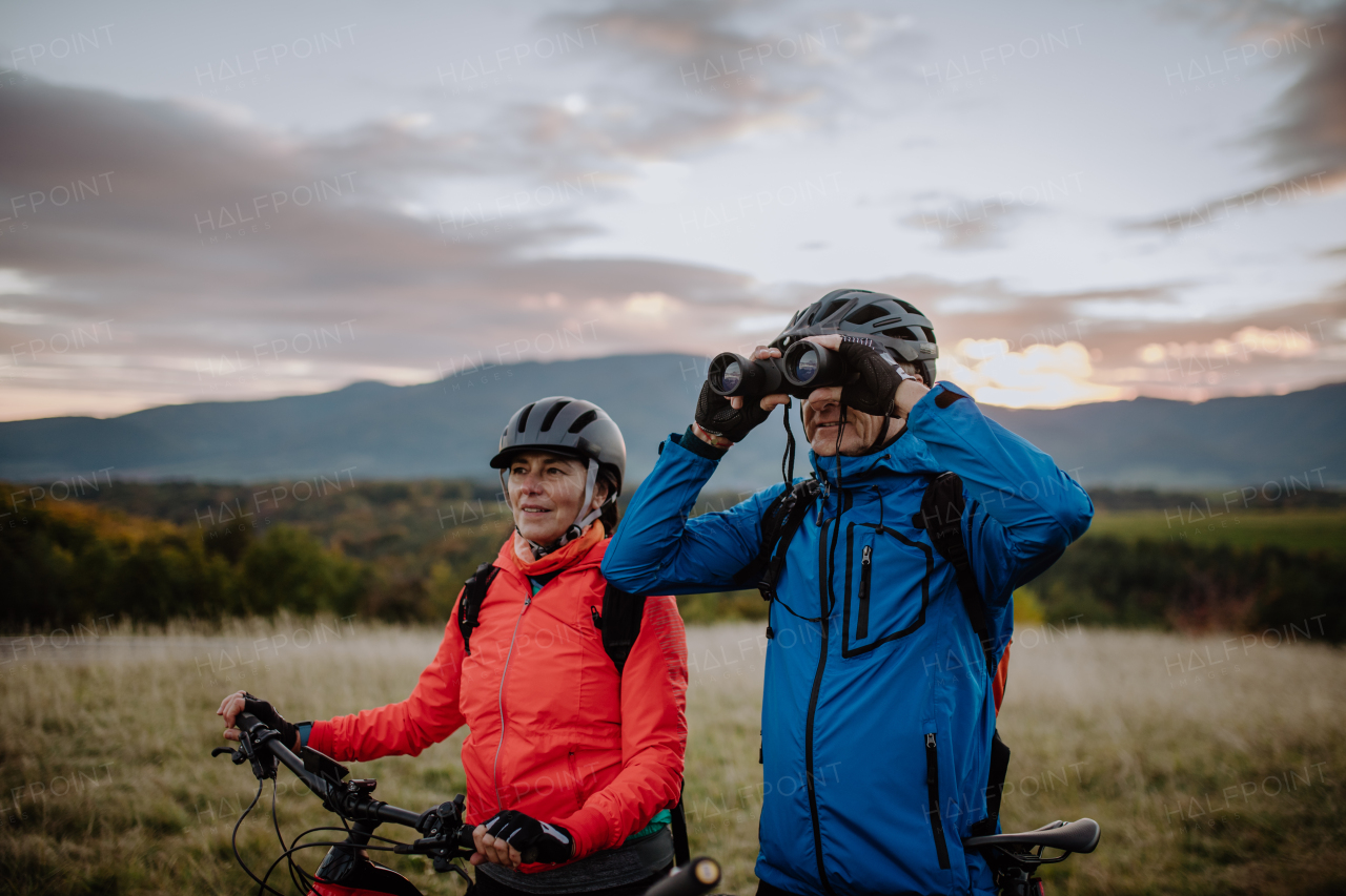 A senior couple bikers with binoculars admiring nature outdoors in meadow in autumn day.