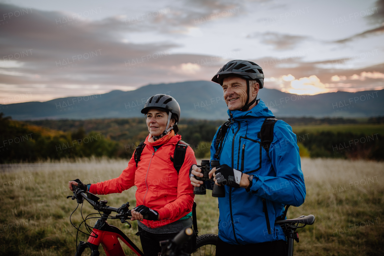 A senior couple bikers with binoculars admiring nature outdoors in meadow in autumn day.