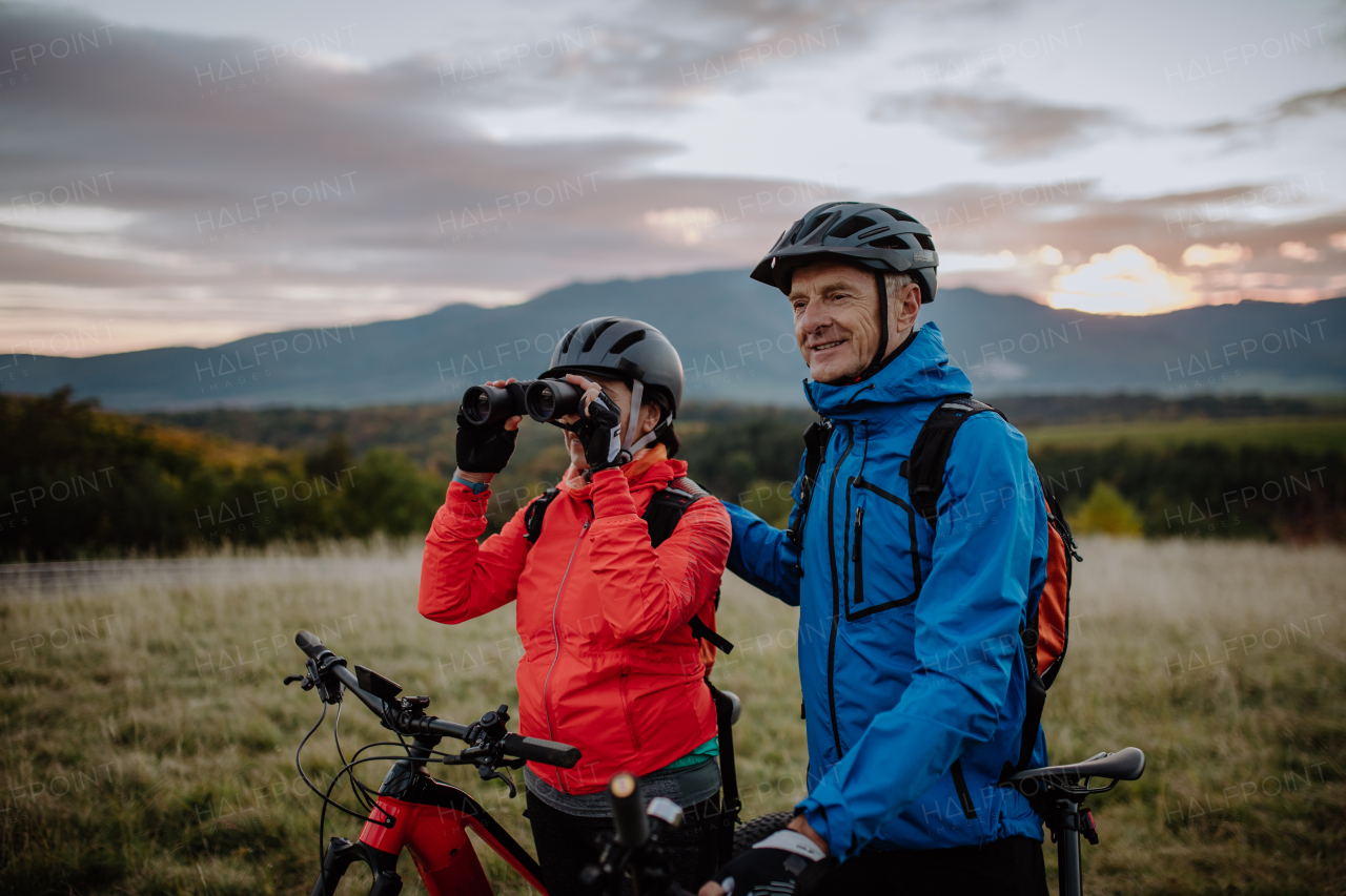 A senior couple bikers with binoculars admiring nature outdoors in meadow in autumn day.