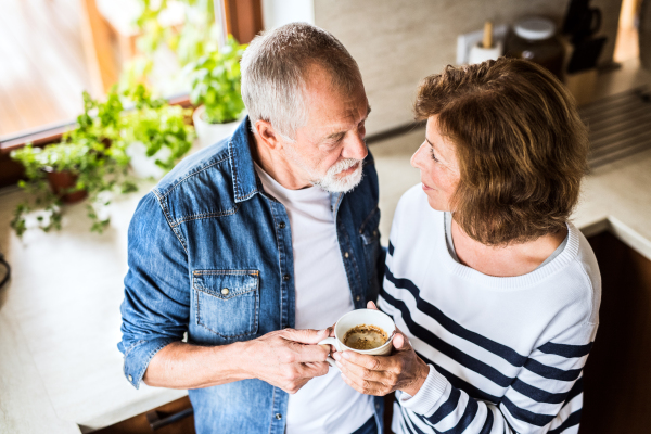 Senior couple talking in the kitchen. An old man and woman inside the house, holding a cup of coffee.