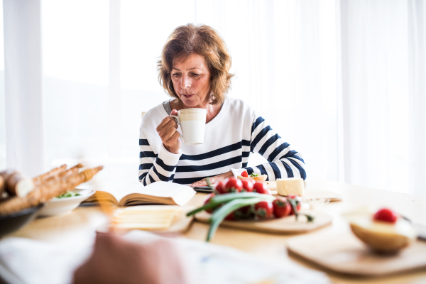 Senior woman eating breakfast at home. An old woman sitting at the table, reading a book.