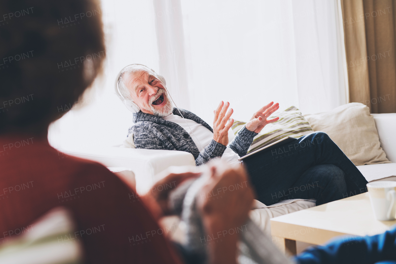 Happy senior couple relaxing at home. An unrecognizable woman knitting and a man with tablet listening to music.