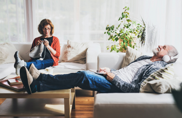 Happy senior couple relaxing at home. A woman knitting and a man with tablet listening to music.