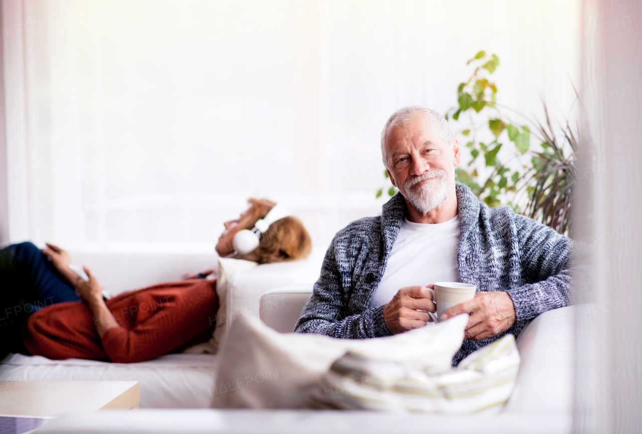 Happy senior couple sitting on the sofa, relaxing at home. A woman listening to music and a man holding a cup of coffee.