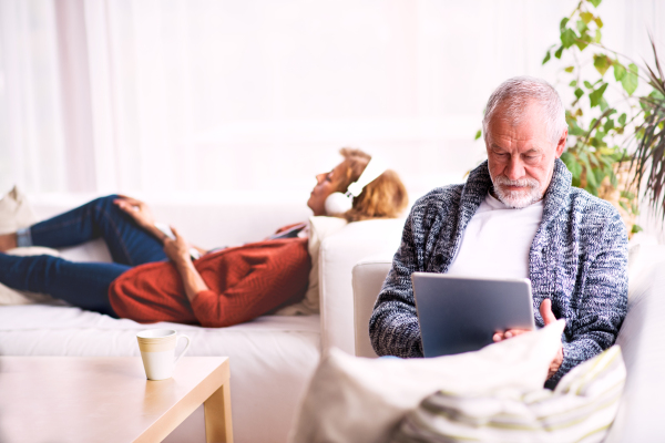 Happy senior couple sitting on the sofa, relaxing at home. A woman listening to music and a man using tablet.