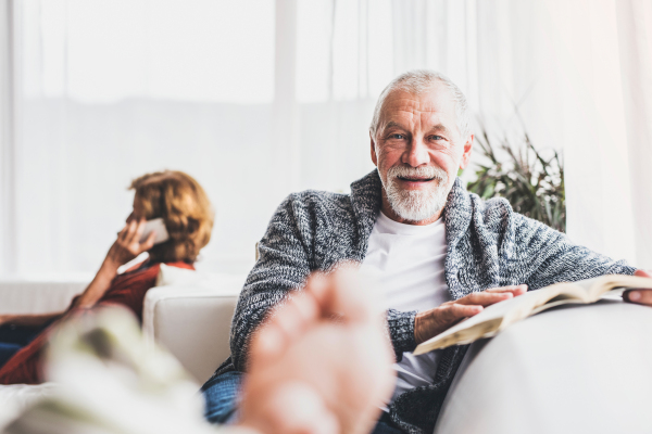 Happy senior couple with smartphone relaxing at home. A woman making a phone call and a man reading a book.