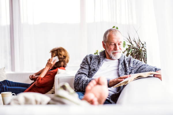 Happy senior couple with smartphone relaxing at home. A woman making a phone call and a man reading a book.