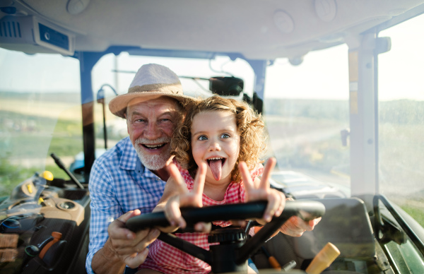 A senior farmer with small granddaughter sitting in tractor, driving.