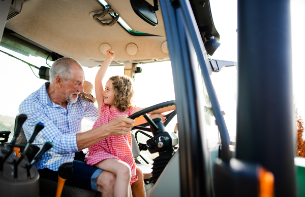 A senior farmer with small granddaughter sitting in tractor, driving.