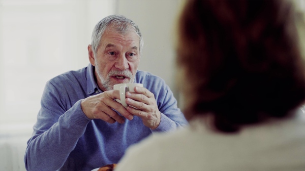 A senior couple with coffee sitting at the table at home, having breakfast and talking.