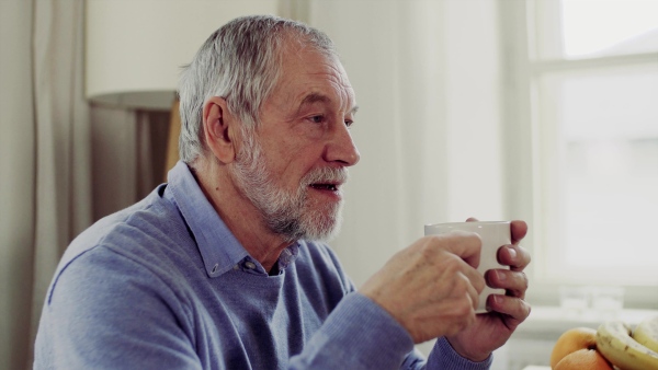 A senior man with a cup of coffee sitting at the table at home, talking to somebody.