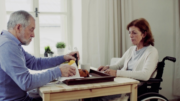 A senior couple with wheelchair sitting at the table at home, having breakfast and talking.