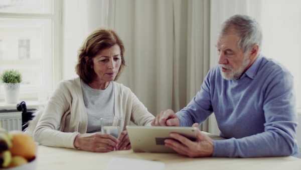 Happy senior couple with wheelchair and tablet sitting at the table at home, talking.