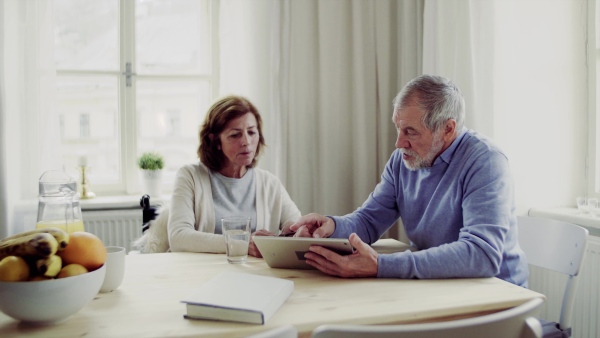 Senior man with tablet explaining his wife in wheelchair how to take medication, sitting at the table.