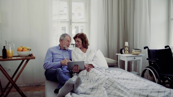 A senior couple with tablet at home, a husband and his wife sitting on bed, spending time together.