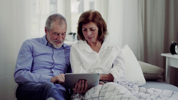 A senior couple with tablet at home, a husband and his wife sitting on bed, spending time together.