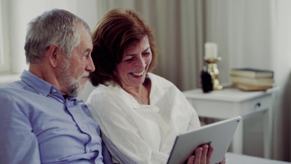 A senior couple with tablet at home, a husband and his wife sitting on bed, spending time together.