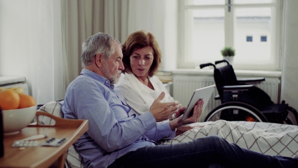 A senior couple with tablet at home, a husband and his wife sitting on bed, spending time together.