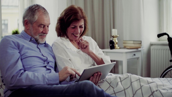 A senior couple with tablet at home, a husband and his wife sitting on bed, spending time together.