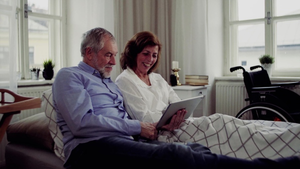 A senior couple with tablet at home, a husband and his wife sitting on bed, spending time together.