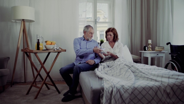 A senior couple sitting on bed at home, a man giving medication to his ill wife.