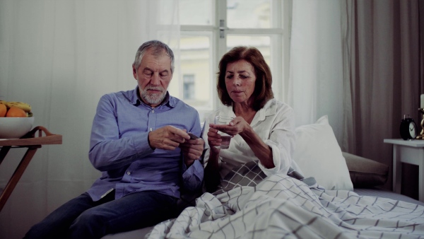 A senior couple sitting on bed at home, a man giving medication to his ill wife.