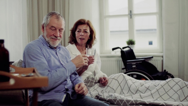 A senior couple sitting on bed at home, a man giving medication to his ill wife.