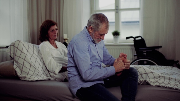 A senior man and his sad disabled wife sitting on bed, giving a foot massage.