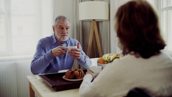 A senior couple with wheelchair sitting at the table at home, having breakfast and talking. Slow motion.