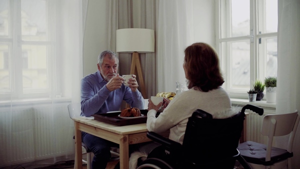 A senior couple with wheelchair sitting at the table at home, having breakfast and talking. Slow motion.