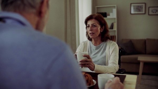 A senior couple with wheelchair sitting at the table at home, having breakfast and talking. Slow motion.