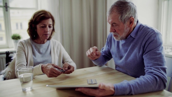 Senior man with tablet explaining his wife in wheelchair how to take medication, sitting at the table. Slow motion.